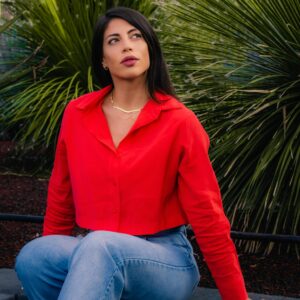Stylish woman in a red top sitting on a bench surrounded by green plants.