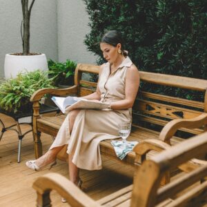 Woman in a beige dress reading a book on a wooden bench in an outdoor patio.