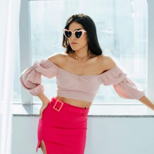 Stylish woman wearing heart-shaped sunglasses and pink attire poses by a sunlit window, exuding confidence and modern fashion.