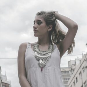 Young woman in a sleeveless white dress and statement necklace poses confidently in a cityscape.